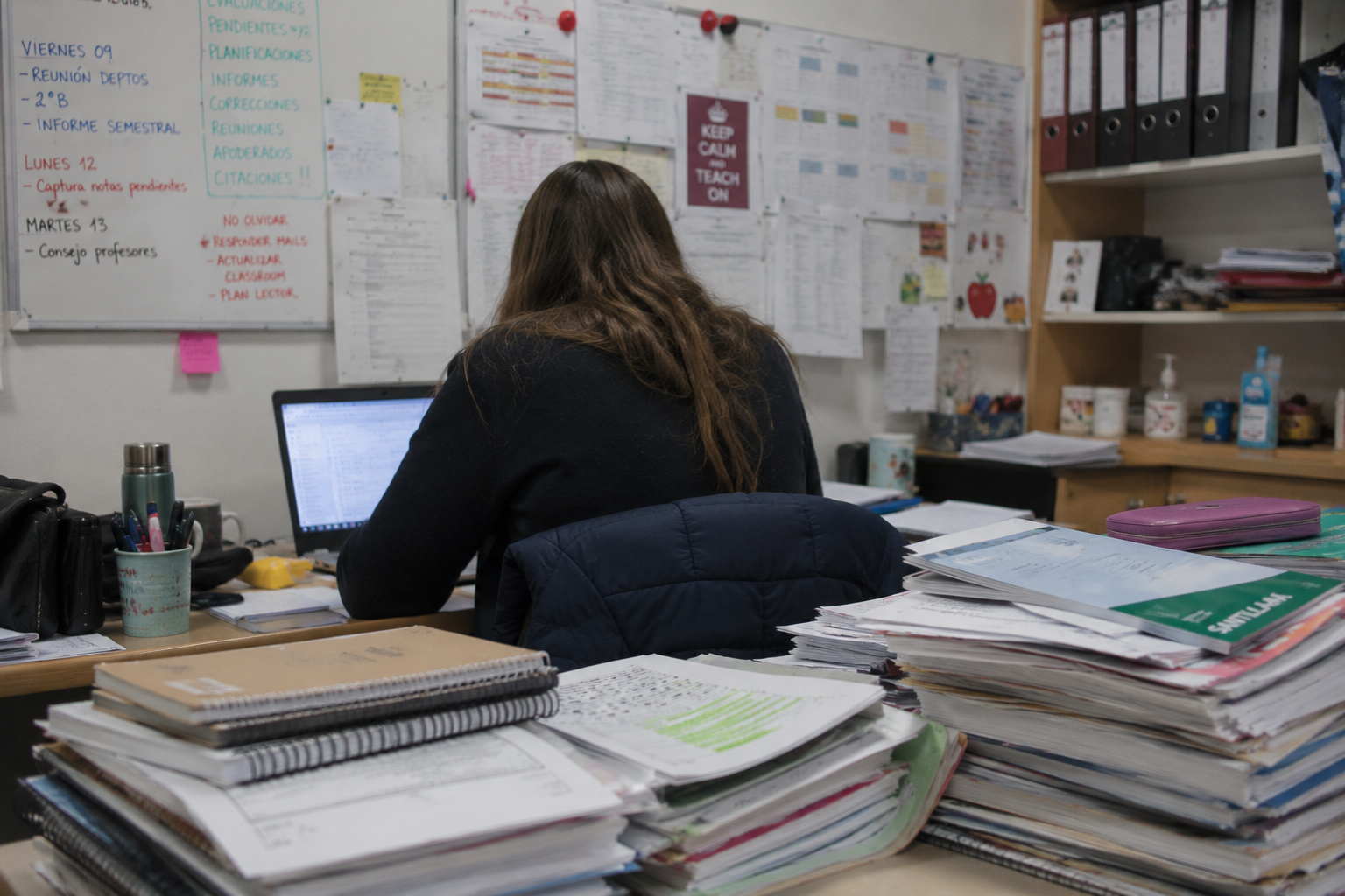 Docente de espaldas trabajando frente a una pizarra llena de tareas pendientes